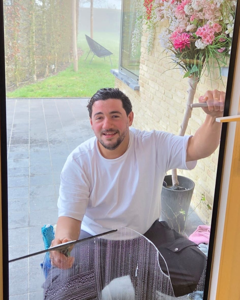 Nabil, zaakvoerder van 4 Seasons Cleaning, in wit T-shirt, lacht door het ingezeepte glas terwijl hij een raam met de wisser schoontrekt; achter hem een terras en bloeiende planten